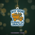 Round-cornered ornament with a blue border and the text Boy Mom Running On Snacks above an orange toy dump truck carrying a teddy bear, a juice cup, a ball and a dinosaur, hanging on a dark green bokeh background.