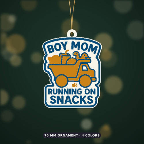 Round-cornered ornament with a blue border and the text Boy Mom Running On Snacks above an orange toy dump truck carrying a teddy bear, a juice cup, a ball and a dinosaur, hanging on a dark green bokeh background.