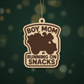 Laser cut wooden ornament engraved with the words Boy Mom Running On Snacks around a brown loaded truck silhouette, hanging on a thin cord against a blurred dark green background.
