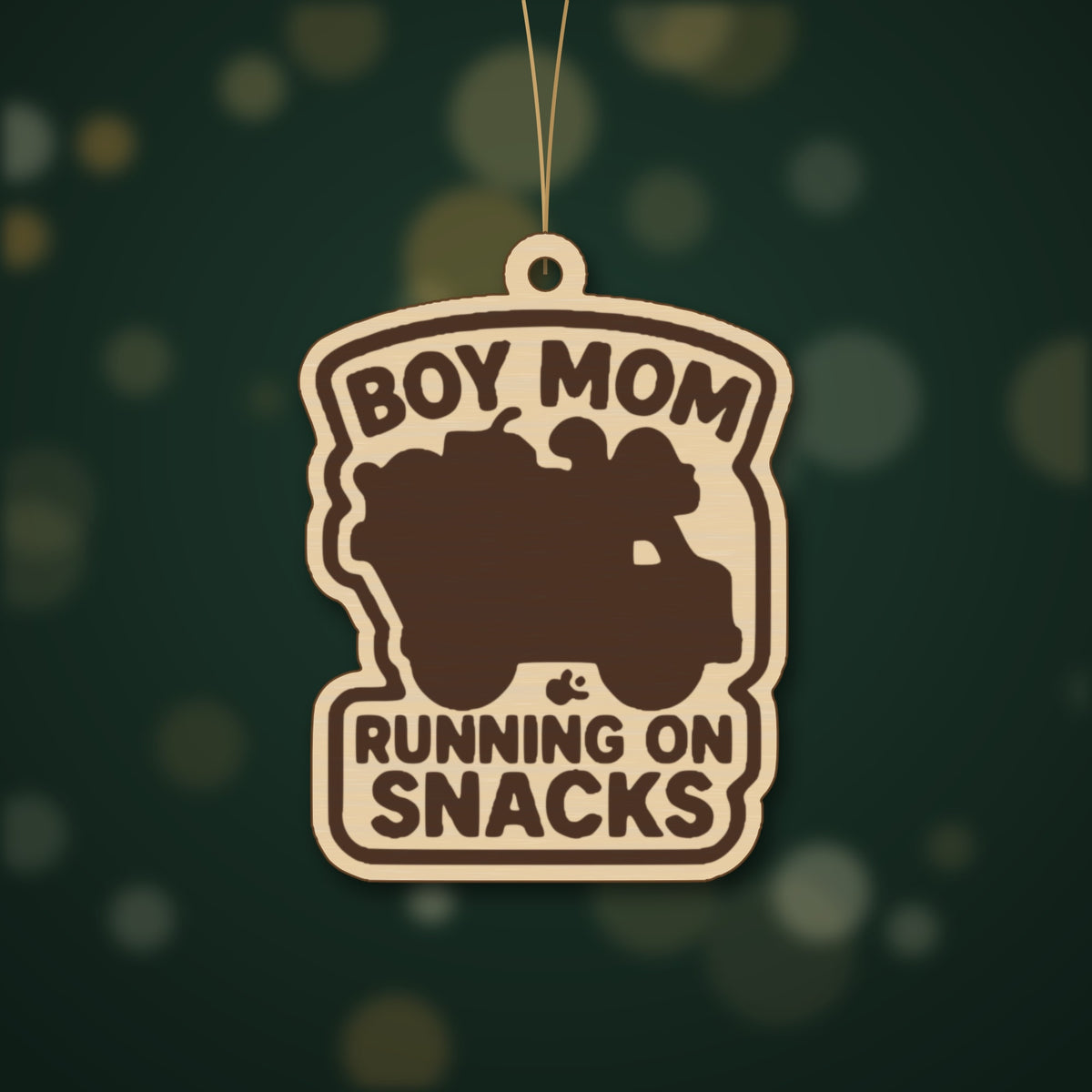 Laser cut wooden ornament engraved with the words Boy Mom Running On Snacks around a brown loaded truck silhouette, hanging on a thin cord against a blurred dark green background.