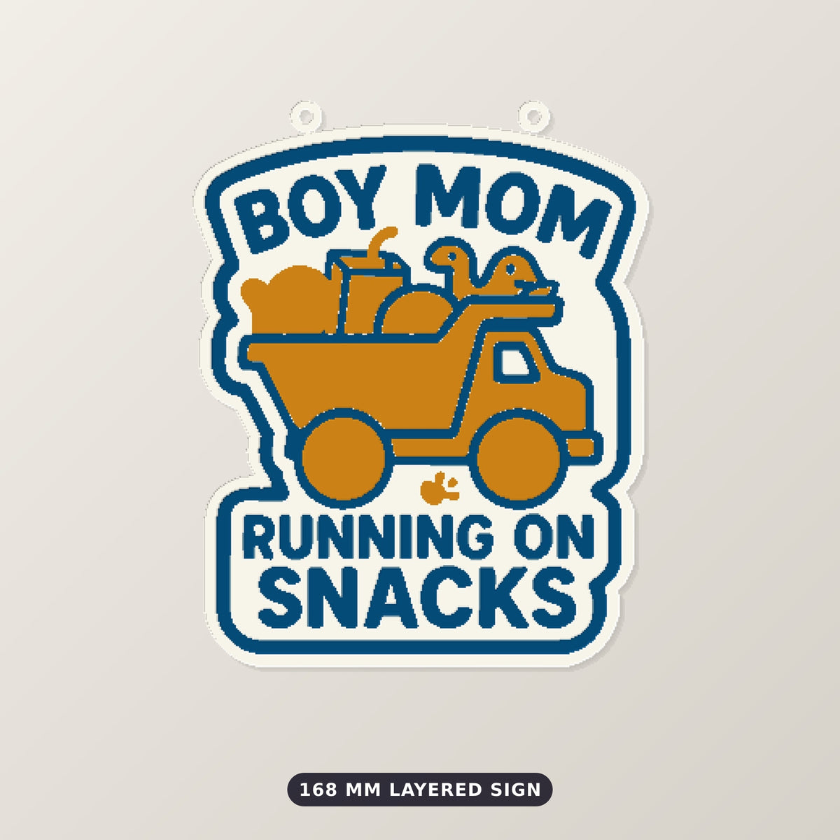 Round-cornered ornament with a blue border and the text Boy Mom Running On Snacks above an orange toy dump truck carrying a teddy bear, a juice cup, a ball and a dinosaur, hanging on a dark green bokeh background.