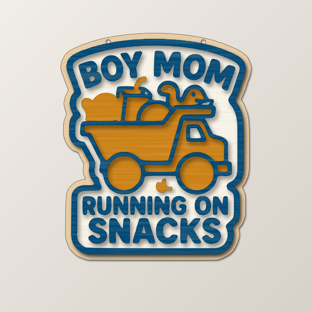 Laser cut wooden ornament engraved with the words Boy Mom Running On Snacks around a brown loaded truck silhouette, hanging on a thin cord against a blurred dark green background.