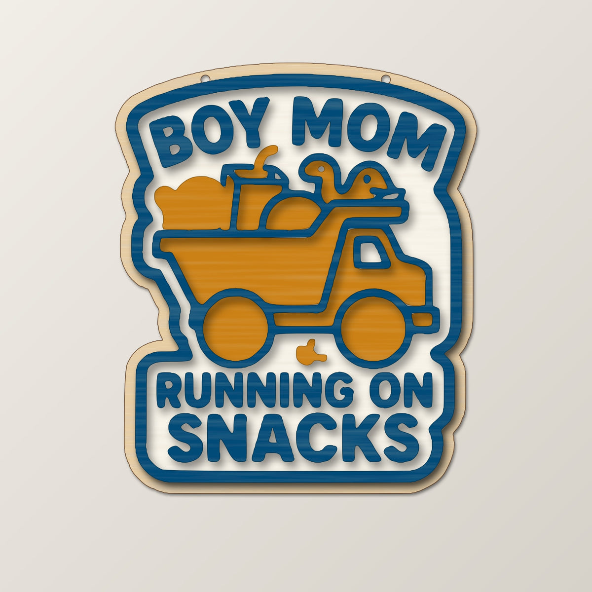 Laser cut wooden ornament engraved with the words Boy Mom Running On Snacks around a brown loaded truck silhouette, hanging on a thin cord against a blurred dark green background.