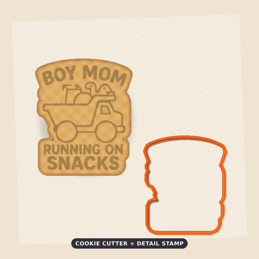 Round-cornered ornament with a blue border and the text Boy Mom Running On Snacks above an orange toy dump truck carrying a teddy bear, a juice cup, a ball and a dinosaur, hanging on a dark green bokeh background.