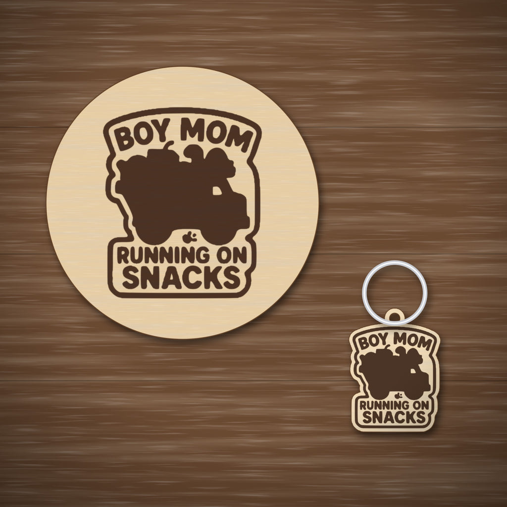 Laser cut wooden ornament engraved with the words Boy Mom Running On Snacks around a brown loaded truck silhouette, hanging on a thin cord against a blurred dark green background.