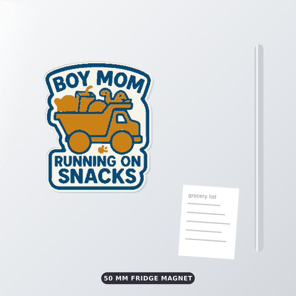 Round-cornered ornament with a blue border and the text Boy Mom Running On Snacks above an orange toy dump truck carrying a teddy bear, a juice cup, a ball and a dinosaur, hanging on a dark green bokeh background.