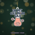 Pixel-style ornament with navy retro wavy text "In My Cat Mom Era" above a pink cat seen from behind with a heart-shaped tail and two flowers, on a dark green bokeh background labeled 75 mm ornament, 4 colors.