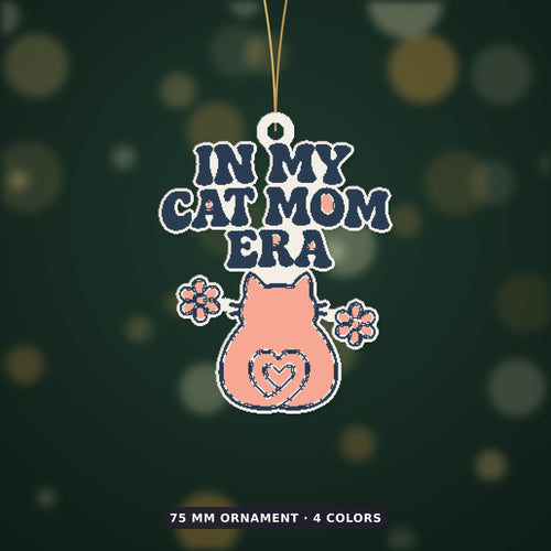 Pixel-style ornament with navy retro wavy text "In My Cat Mom Era" above a pink cat seen from behind with a heart-shaped tail and two flowers, on a dark green bokeh background labeled 75 mm ornament, 4 colors.