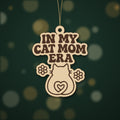 Laser cut wooden ornament engraved with the retro wavy text In My Cat Mom Era, a cat silhouette with a heart-shaped tail and two flowers, hanging on a dark green bokeh background.
