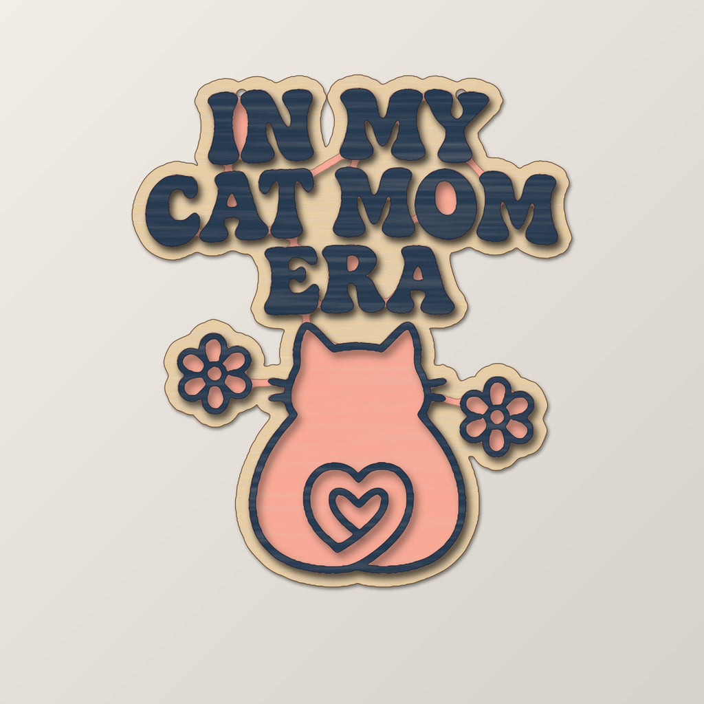 Laser cut wooden ornament engraved with the retro wavy text In My Cat Mom Era, a cat silhouette with a heart-shaped tail and two flowers, hanging on a dark green bokeh background.