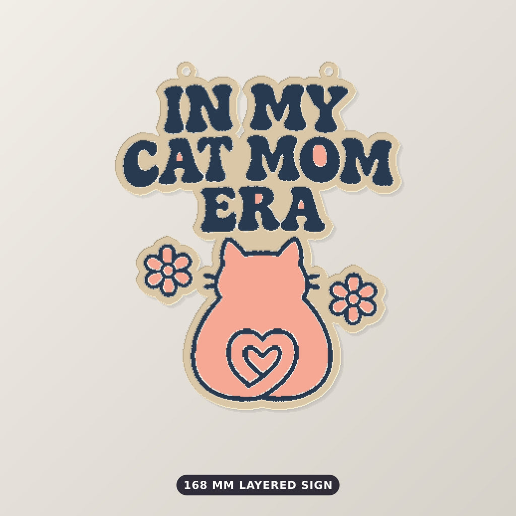 Pixel-style ornament with navy retro wavy text "In My Cat Mom Era" above a pink cat seen from behind with a heart-shaped tail and two flowers, on a dark green bokeh background labeled 75 mm ornament, 4 colors.