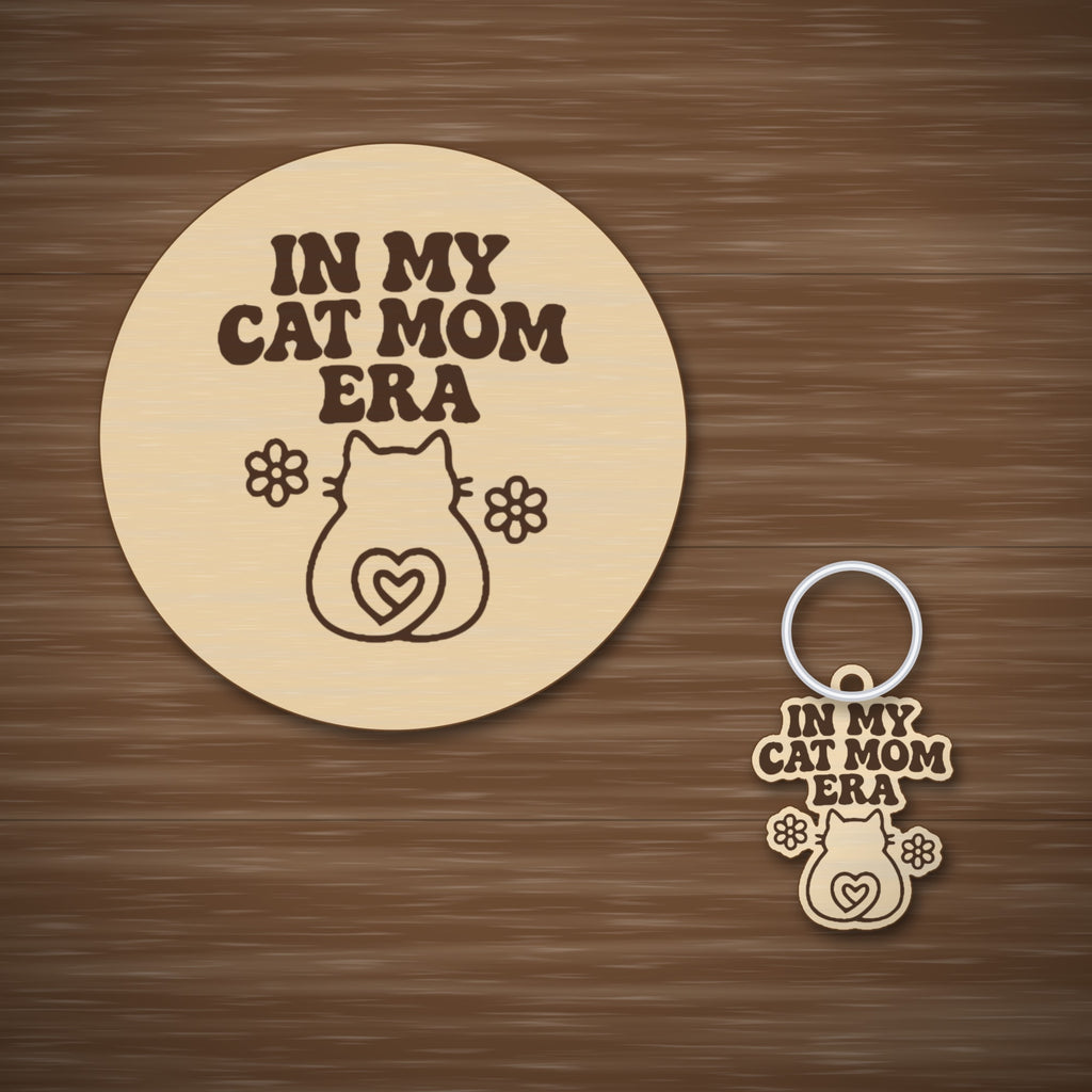 Laser cut wooden ornament engraved with the retro wavy text In My Cat Mom Era, a cat silhouette with a heart-shaped tail and two flowers, hanging on a dark green bokeh background.