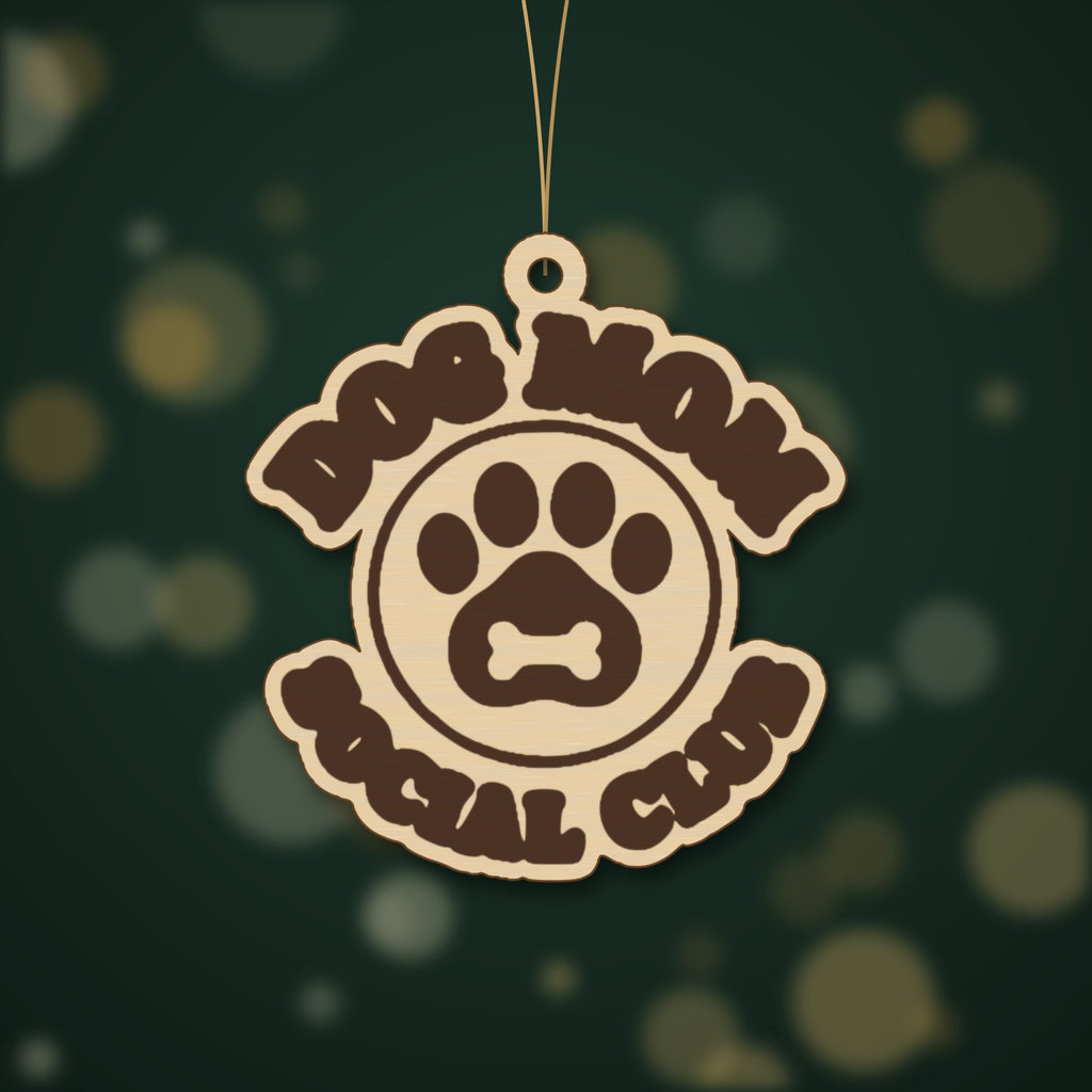 Round wooden laser cut ornament engraved with the words Dog Mom Social Club around a paw print with a bone, hanging on a thin cord against a dark green background with golden bokeh lights.