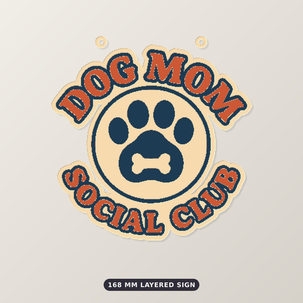 Round Dog Mom Social Club ornament with an orange retro lettering border and a navy paw print with a bone on a cream badge, hanging on a gold string against a dark green background with golden bokeh lights.