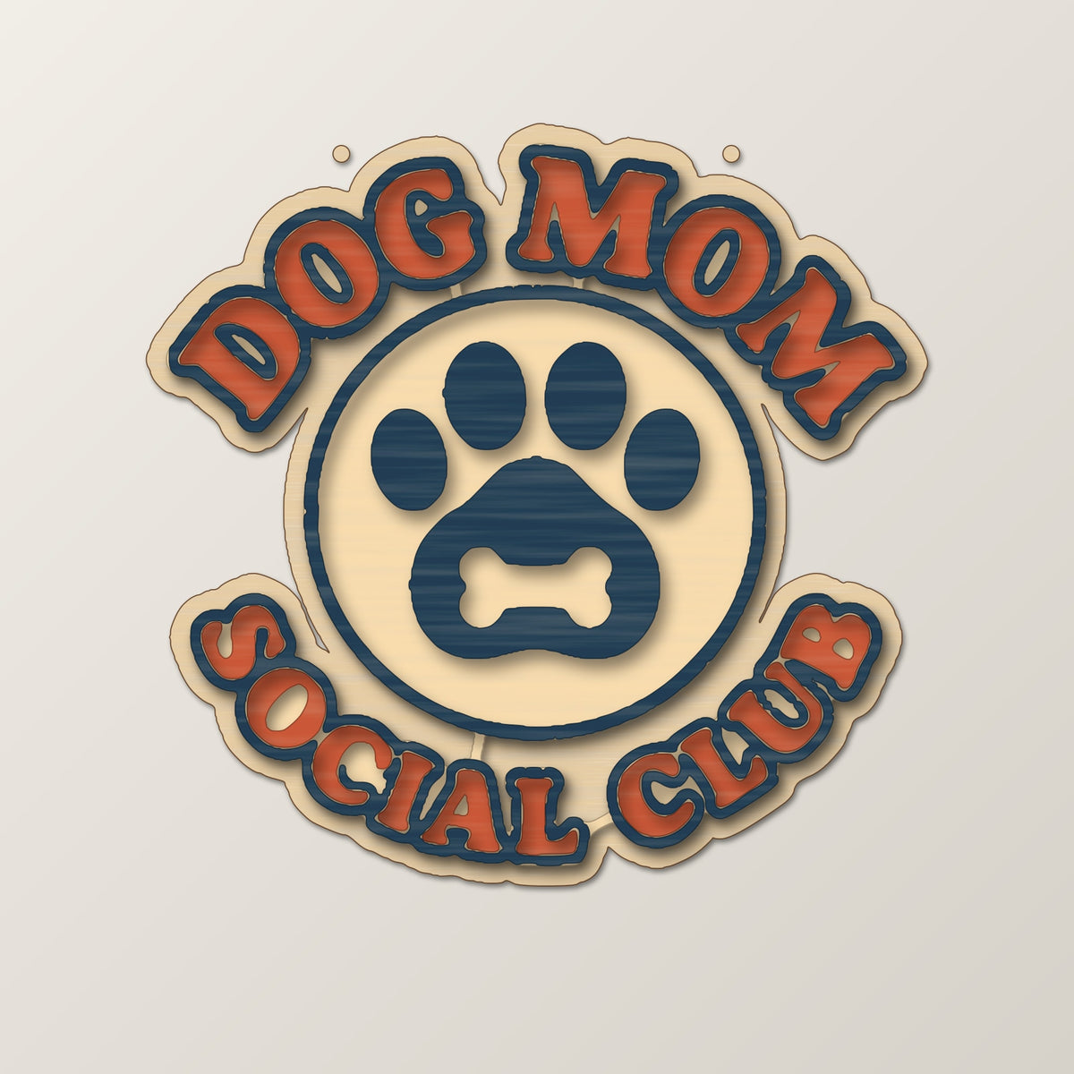 Round wooden laser cut ornament engraved with the words Dog Mom Social Club around a paw print with a bone, hanging on a thin cord against a dark green background with golden bokeh lights.