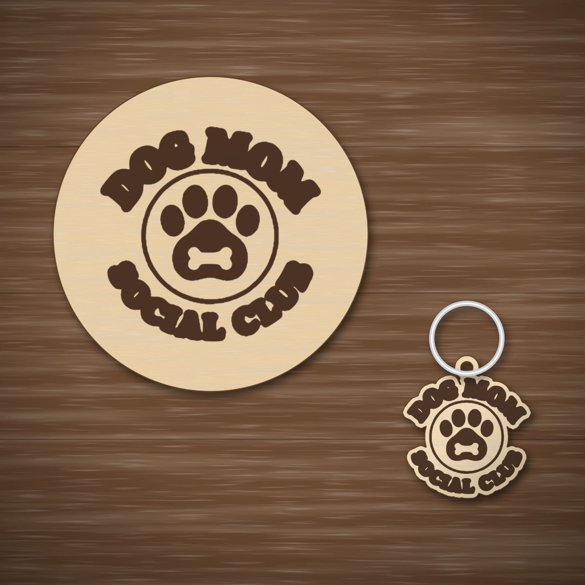 Round wooden laser cut ornament engraved with the words Dog Mom Social Club around a paw print with a bone, hanging on a thin cord against a dark green background with golden bokeh lights.