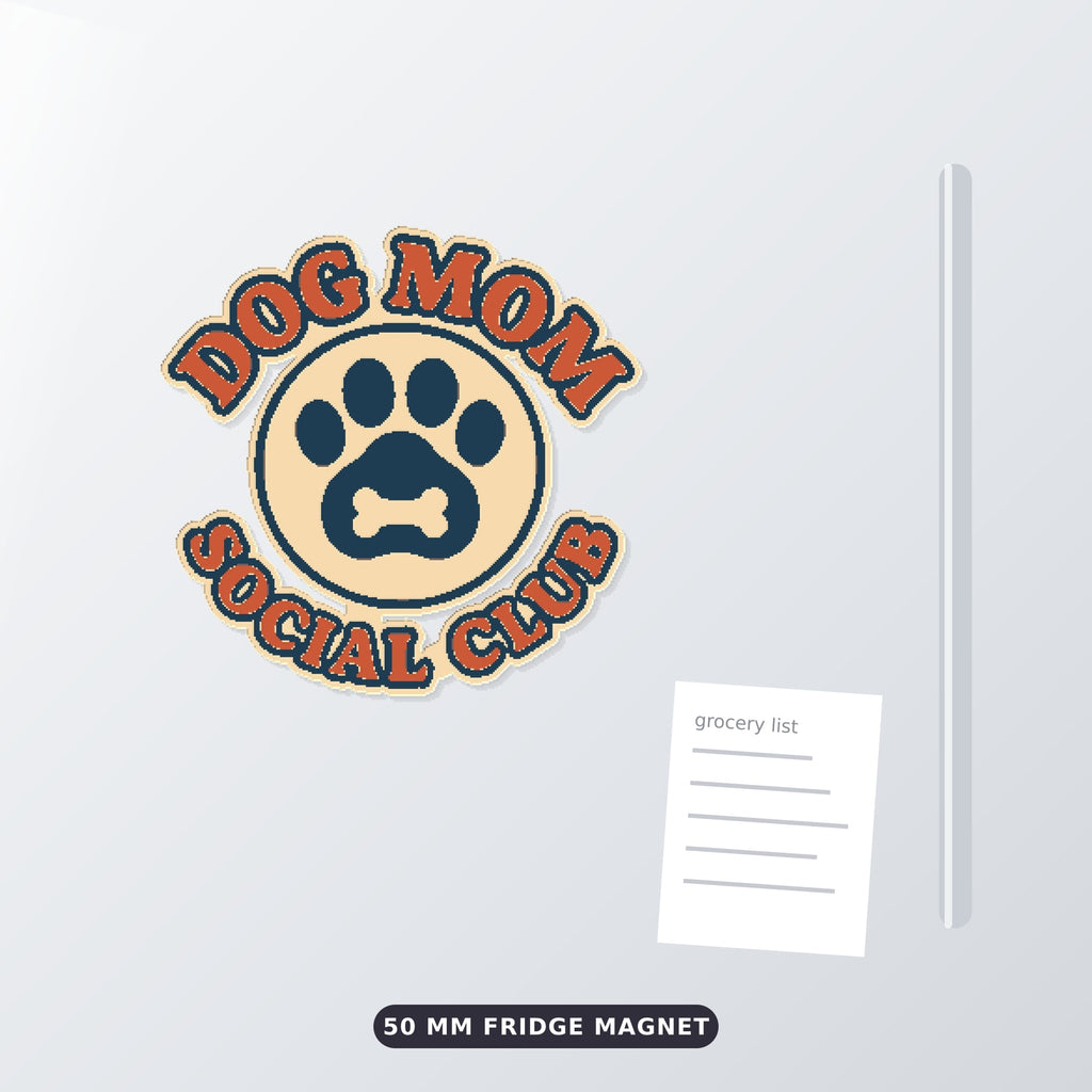 Round Dog Mom Social Club ornament with an orange retro lettering border and a navy paw print with a bone on a cream badge, hanging on a gold string against a dark green background with golden bokeh lights.