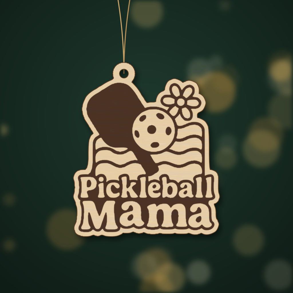 A wooden laser cut ornament with a groovy engraved pickleball paddle, ball, daisy and wavy lines above the words Pickleball Mama, hanging on a dark green bokeh background.