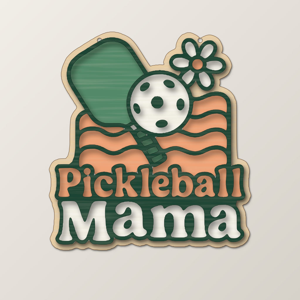 A wooden laser cut ornament with a groovy engraved pickleball paddle, ball, daisy and wavy lines above the words Pickleball Mama, hanging on a dark green bokeh background.
