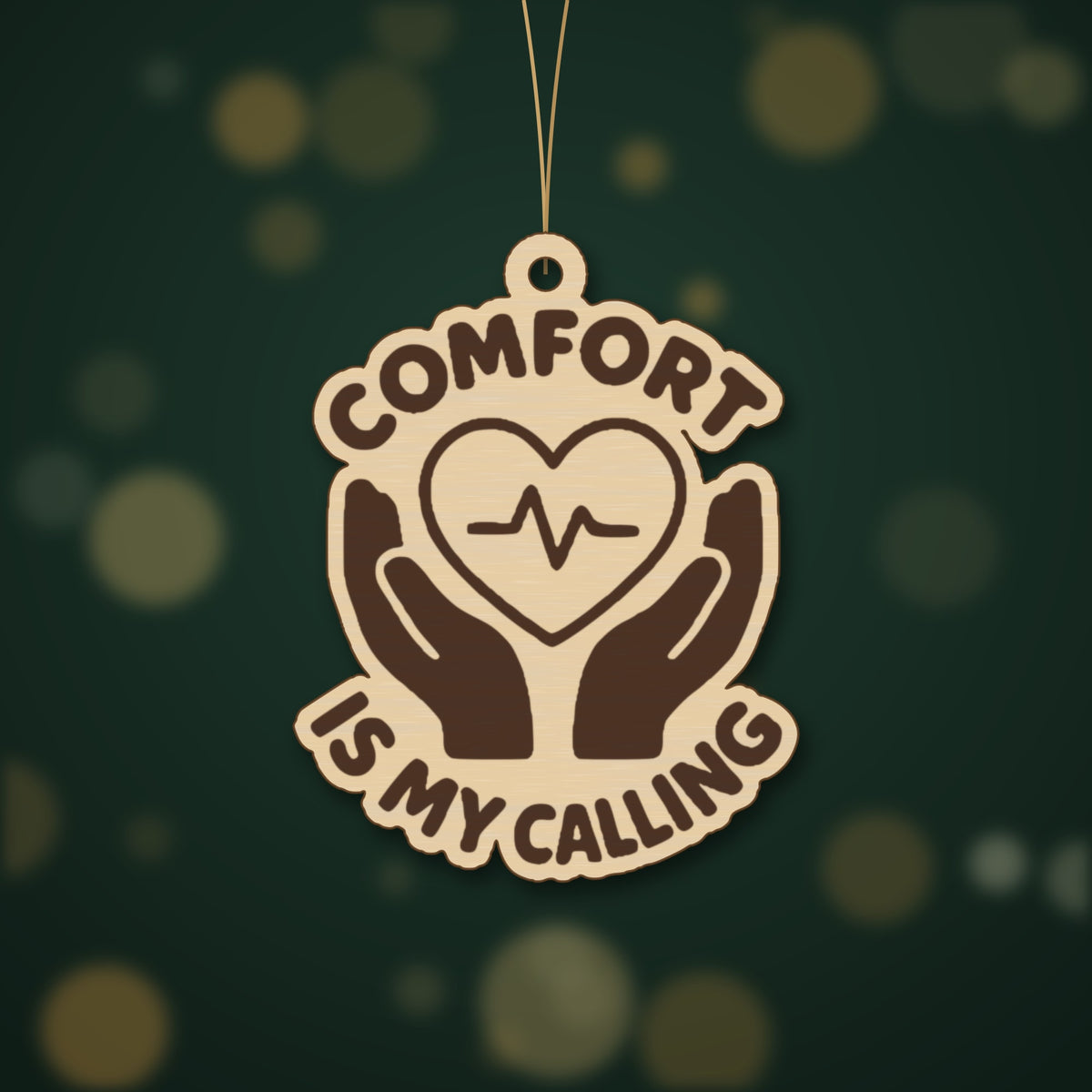 Round-edged wooden ornament with an engraved heart and pulse line held by two hands, with the words Comfort Is My Calling, hanging against a dark green blurred light background.