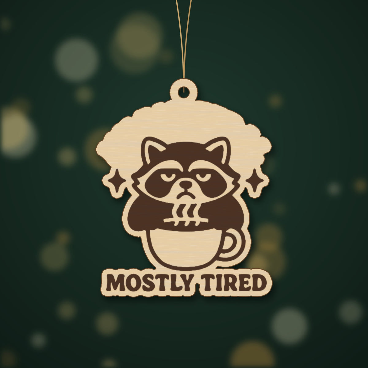 A laser-cut wooden ornament showing a grumpy raccoon sitting in a coffee cup with sparkles above it and the words Mostly Tired engraved underneath, hanging on a gold string against a dark green background.