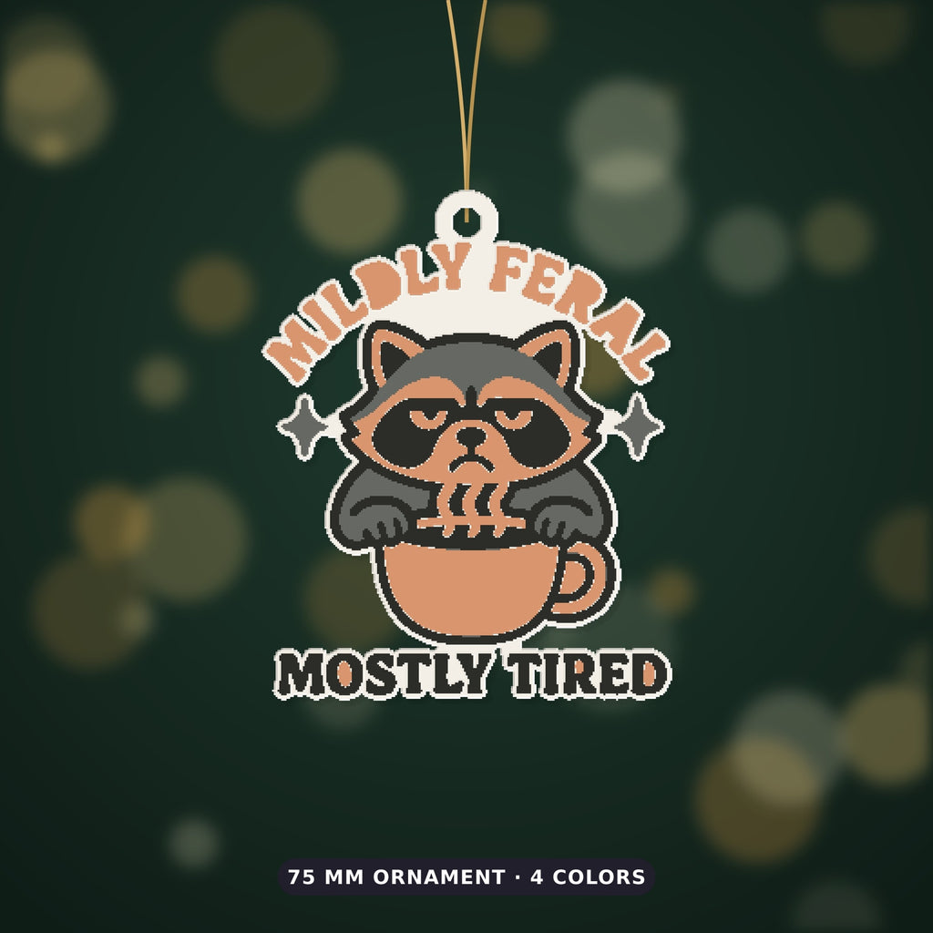 Grumpy raccoon peeking over a coffee cup on an ornament with the text Mildly Feral Mostly Tired, shown on a dark green background.