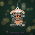 Grumpy raccoon peeking over a coffee cup on an ornament with the text Mildly Feral Mostly Tired, shown on a dark green background.