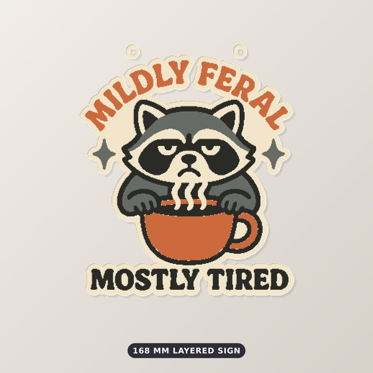 Grumpy raccoon peeking over a coffee cup on an ornament with the text Mildly Feral Mostly Tired, shown on a dark green background.