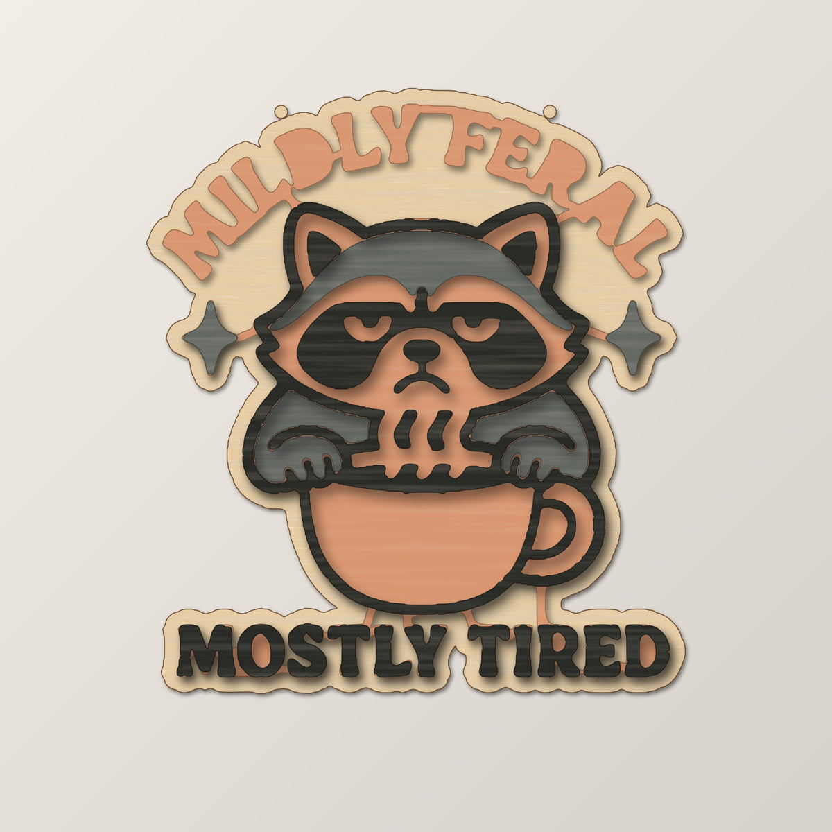 A laser-cut wooden ornament showing a grumpy raccoon sitting in a coffee cup with sparkles above it and the words Mostly Tired engraved underneath, hanging on a gold string against a dark green background.