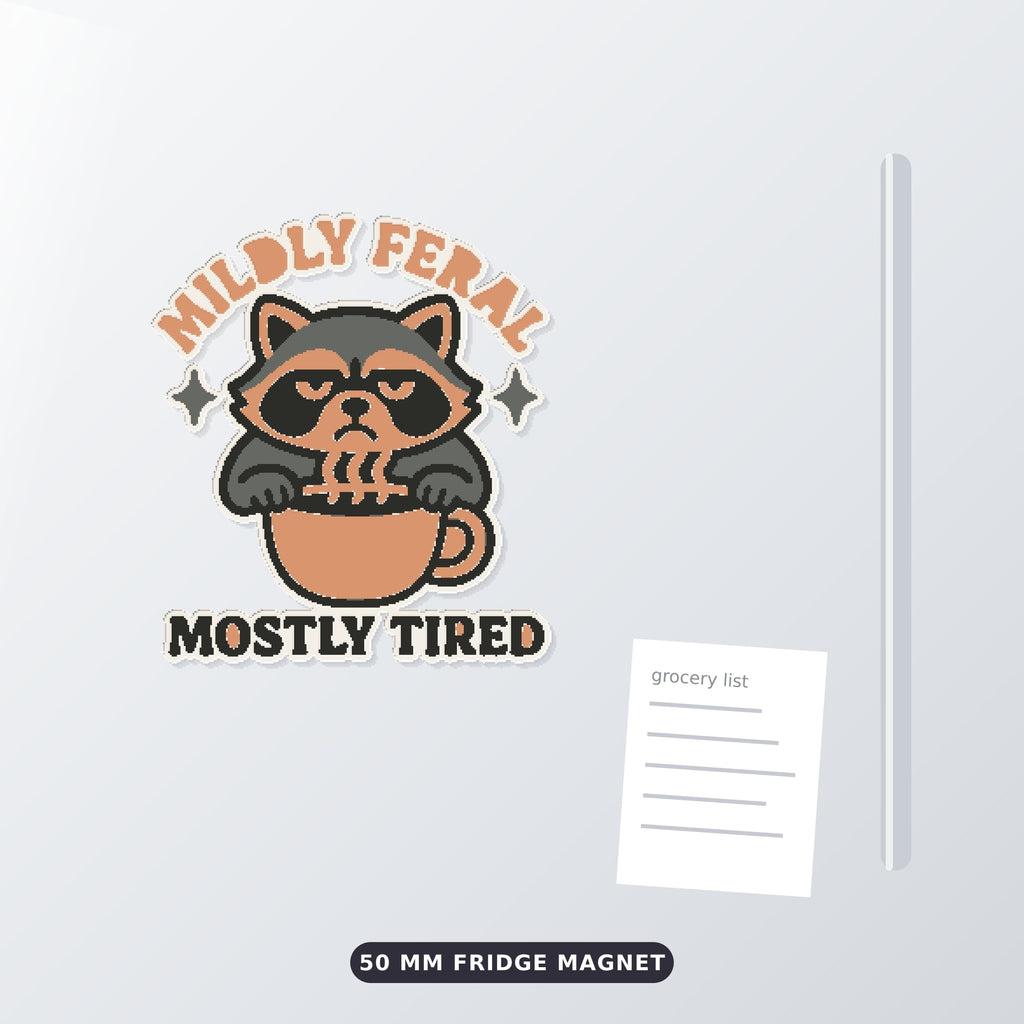 Grumpy raccoon peeking over a coffee cup on an ornament with the text Mildly Feral Mostly Tired, shown on a dark green background.