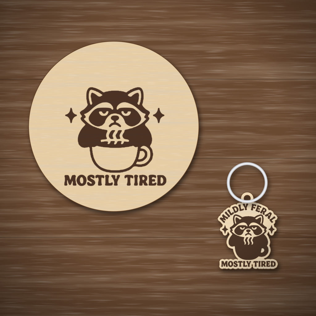 A laser-cut wooden ornament showing a grumpy raccoon sitting in a coffee cup with sparkles above it and the words Mostly Tired engraved underneath, hanging on a gold string against a dark green background.