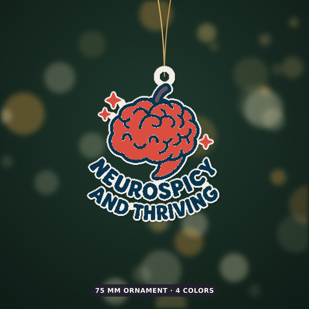 A 75 mm ornament of a smiling red chili-pepper brain with sparkles and the words Neurospicy and Thriving, hanging on a gold cord against a dark green bokeh background.