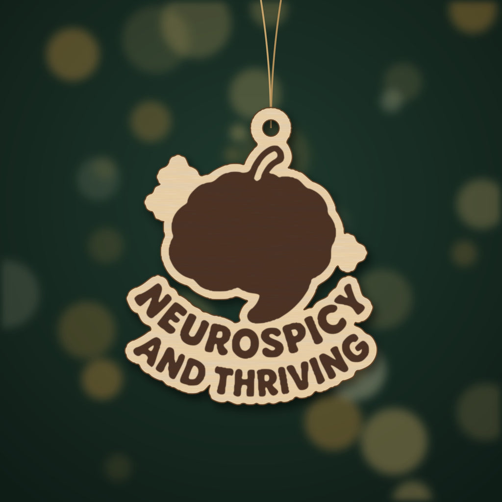 Wooden laser cut ornament with a dark brown engraved brain with a chili stem and the words Neurospicy and Thriving, hanging on a gold string against a green background with soft bokeh lights.