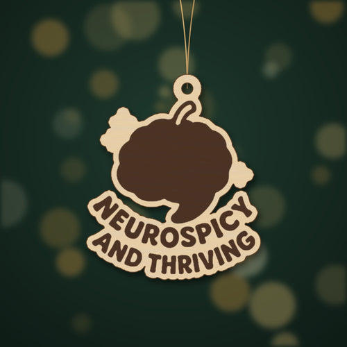 Wooden laser cut ornament with a dark brown engraved brain with a chili stem and the words Neurospicy and Thriving, hanging on a gold string against a green background with soft bokeh lights.