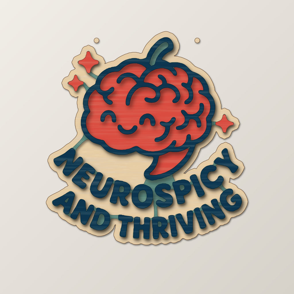 Wooden laser cut ornament with a dark brown engraved brain with a chili stem and the words Neurospicy and Thriving, hanging on a gold string against a green background with soft bokeh lights.