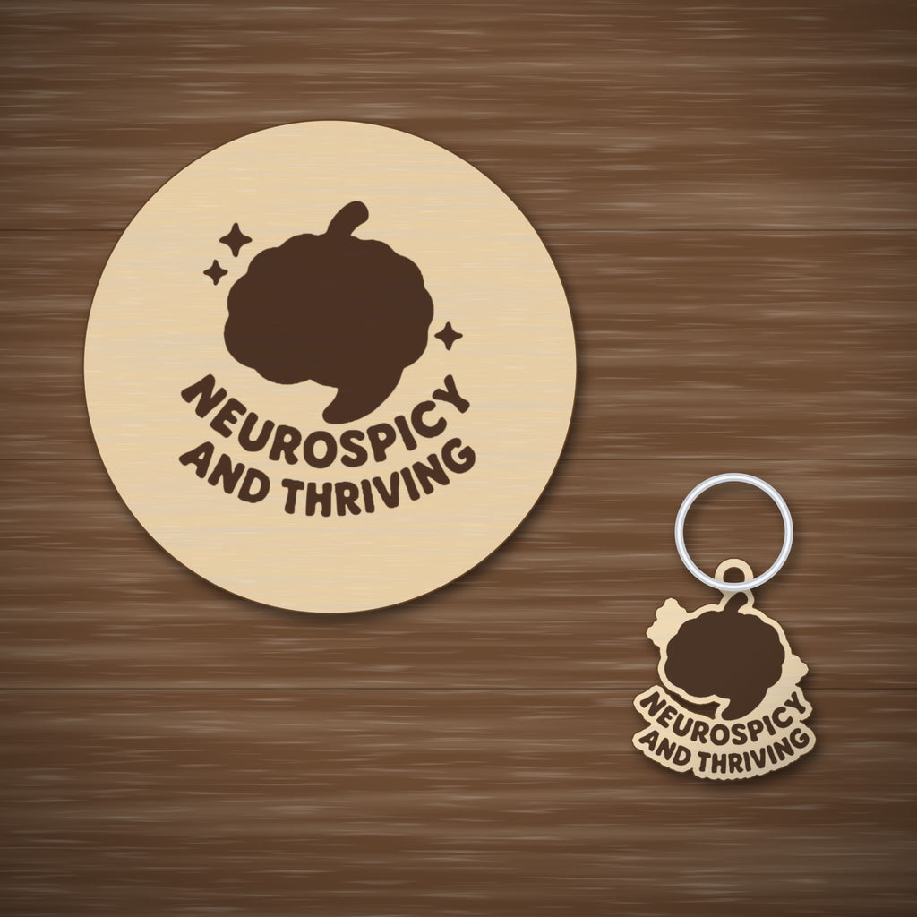 Wooden laser cut ornament with a dark brown engraved brain with a chili stem and the words Neurospicy and Thriving, hanging on a gold string against a green background with soft bokeh lights.