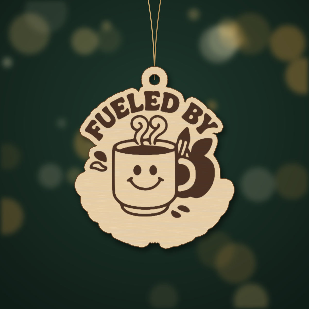 Round wooden laser-engraved ornament with the words Fueled By above a smiling coffee mug, a pencil and an apple, hanging on a thin cord against a dark green background with golden bokeh lights.