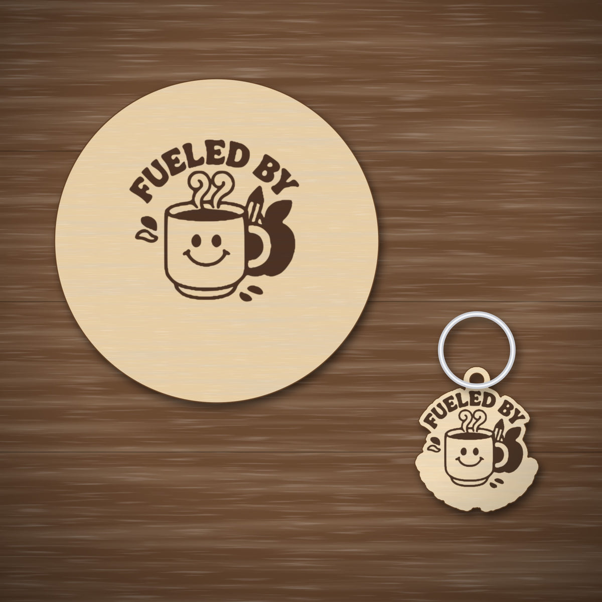 Round wooden laser-engraved ornament with the words Fueled By above a smiling coffee mug, a pencil and an apple, hanging on a thin cord against a dark green background with golden bokeh lights.