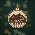 Round wooden laser cut ornament showing a brown bear walking in front of mountains above the words Mama Bear, hanging on a thin cord against a blurred green background with golden lights.