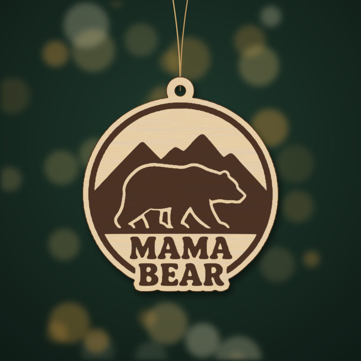 Round wooden laser cut ornament showing a brown bear walking in front of mountains above the words Mama Bear, hanging on a thin cord against a blurred green background with golden lights.
