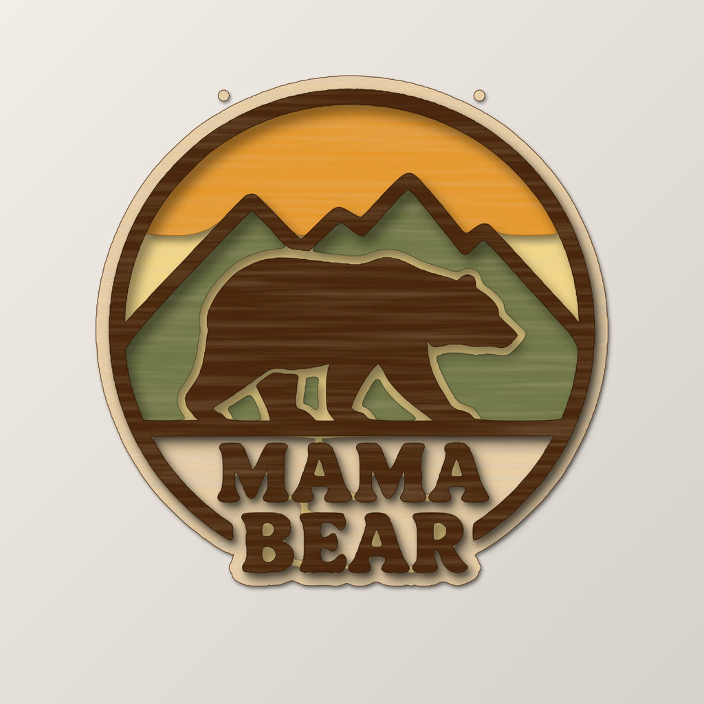 Round wooden laser cut ornament showing a brown bear walking in front of mountains above the words Mama Bear, hanging on a thin cord against a blurred green background with golden lights.