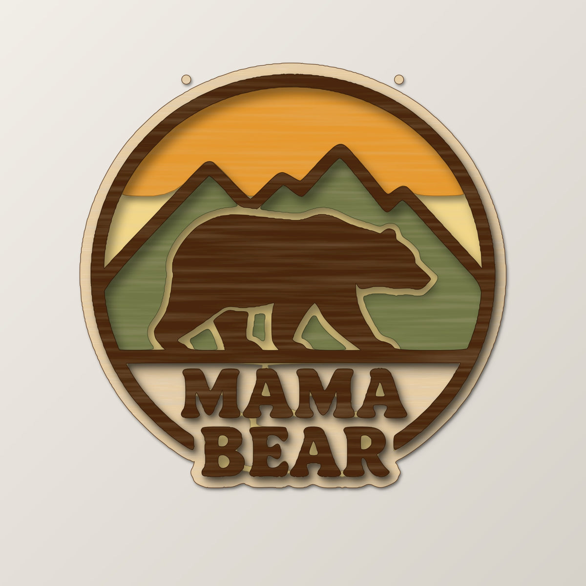 Round wooden laser cut ornament showing a brown bear walking in front of mountains above the words Mama Bear, hanging on a thin cord against a blurred green background with golden lights.