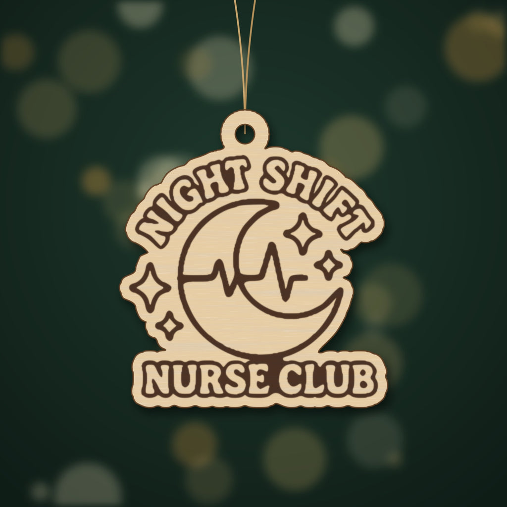 A round-cornered wooden laser cut ornament engraved with a crescent moon, heartbeat line and sparkles, with the words Night Shift Nurse Club, hanging on a gold string against a dark green background with soft lights.