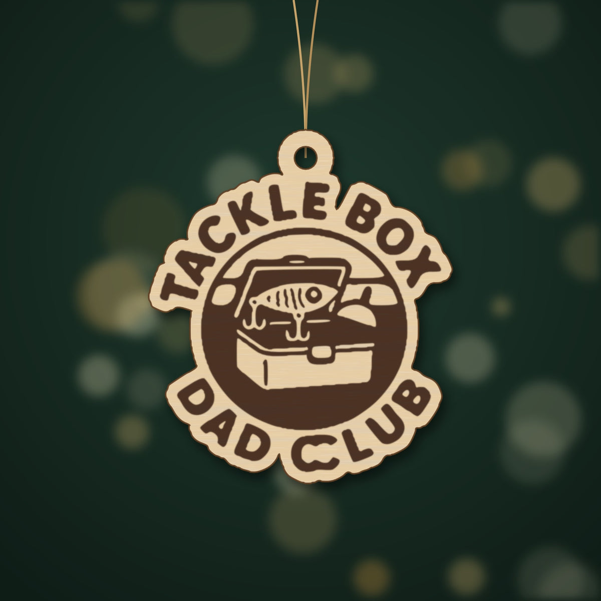 Round wooden laser engraved ornament with a retro tackle box and fishing lure badge and the words Tackle Box Dad Club, hanging on a thin cord against a dark green background with soft golden lights.