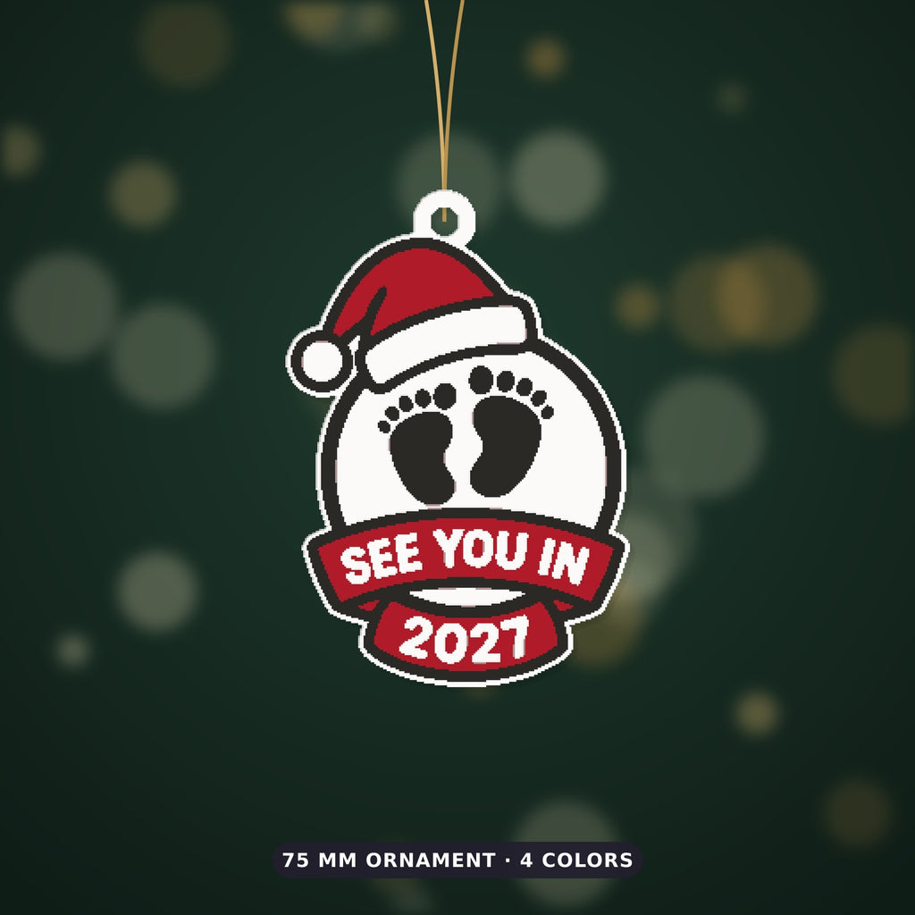 Round white ornament with black baby footprints, a red Santa hat and a red ribbon banner reading See You In 2027, hanging on a gold string against a dark green background.
