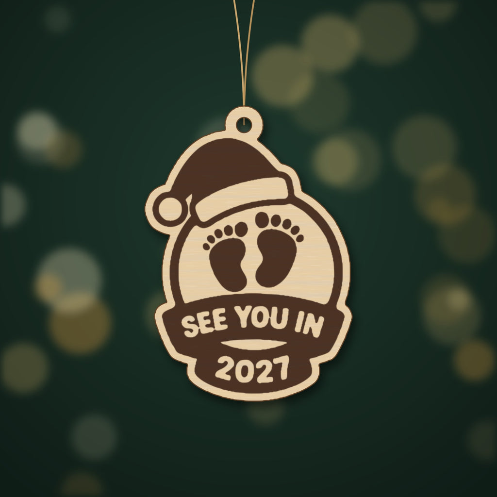 Wooden laser cut Christmas ornament with a Santa hat, engraved baby footprints and a banner reading See You In 2027, hanging against a blurred green background with gold lights.
