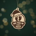 Wooden laser cut Christmas ornament with a Santa hat, engraved baby footprints and a banner reading See You In 2027, hanging against a blurred green background with gold lights.