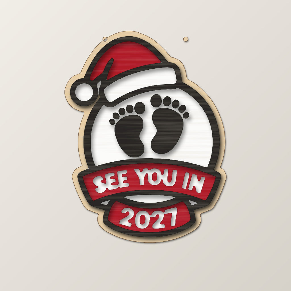 Wooden laser cut Christmas ornament with a Santa hat, engraved baby footprints and a banner reading See You In 2027, hanging against a blurred green background with gold lights.