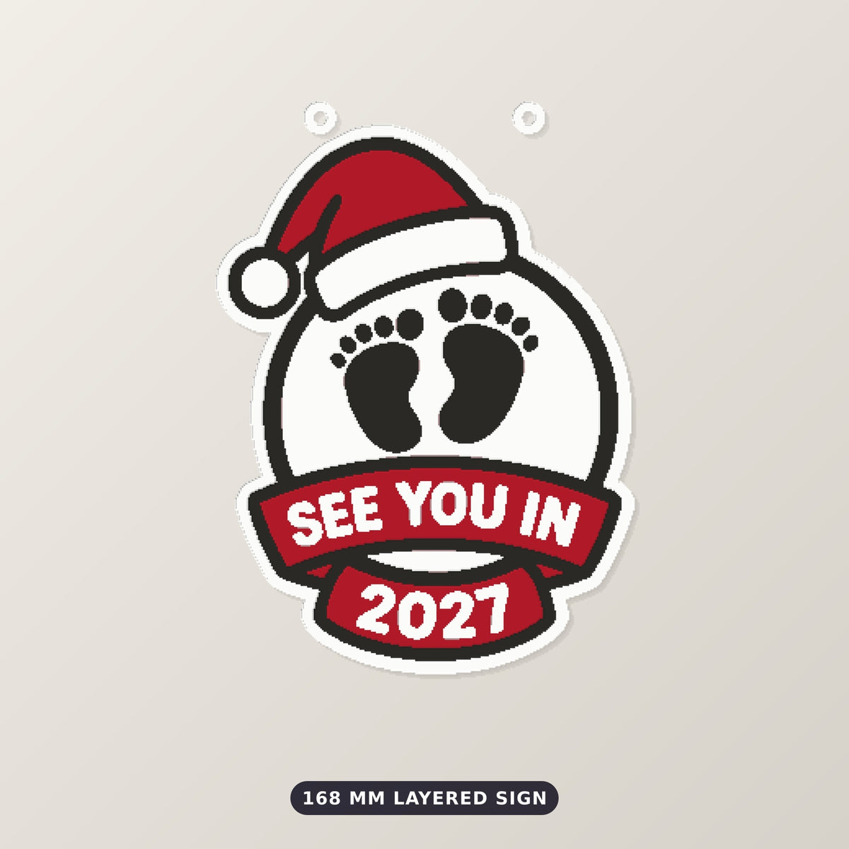 Round white ornament with black baby footprints, a red Santa hat and a red ribbon banner reading See You In 2027, hanging on a gold string against a dark green background.