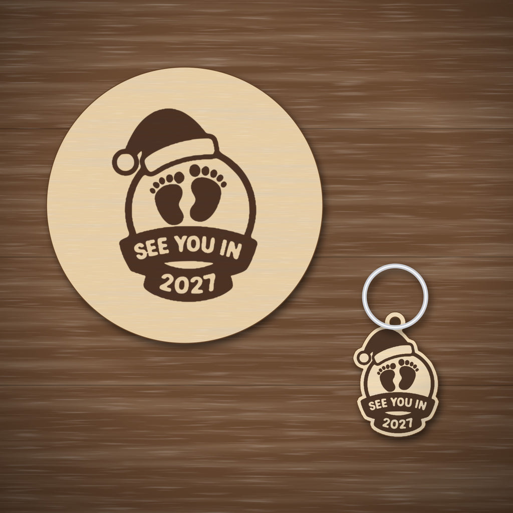 Wooden laser cut Christmas ornament with a Santa hat, engraved baby footprints and a banner reading See You In 2027, hanging against a blurred green background with gold lights.